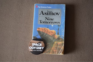 A vintage science fiction book titled 'Nine Tomorrows' by Isaac Asimov is placed on a textured surface. The book features a colorful cover with an abstract illustration. In front of the book, there is a round badge with text that reads 'Space Odyssey' and 'Fear is the mind-killer'. The overall image has a nostalgic and sci-fi theme.