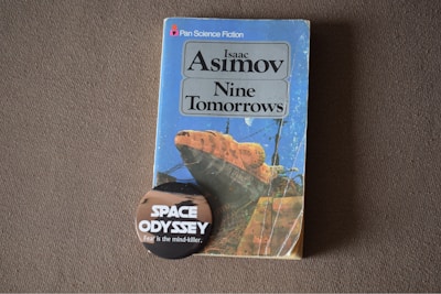 A vintage science fiction book titled 'Nine Tomorrows' by Isaac Asimov is placed on a textured surface. The book features a colorful cover with an abstract illustration. In front of the book, there is a round badge with text that reads 'Space Odyssey' and 'Fear is the mind-killer'. The overall image has a nostalgic and sci-fi theme.