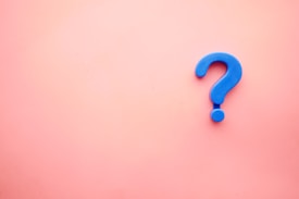 A large, blue plastic question mark is centered against a smooth, pink background.