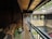A wooden balcony corridor is decorated with a wicker chair and a potted plant in the corner. The architecture features traditional wooden railings and beams. Across the courtyard, brick walls with intricate wooden detailing frame potted plants hanging on the wall.