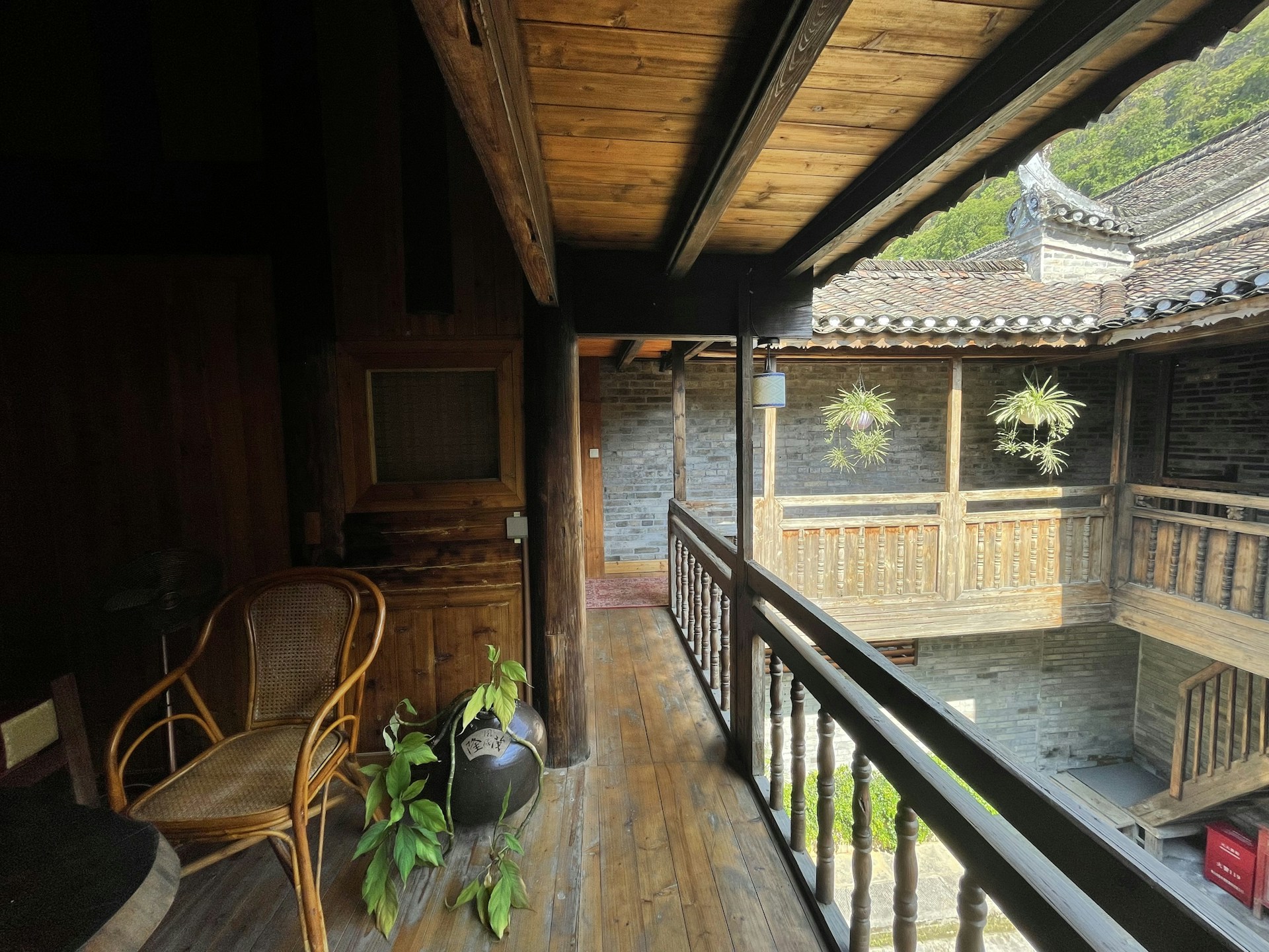 A wooden balcony corridor is decorated with a wicker chair and a potted plant in the corner. The architecture features traditional wooden railings and beams. Across the courtyard, brick walls with intricate wooden detailing frame potted plants hanging on the wall.