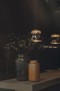 Two decorative vases, one dark green and the other orange, sit on a wooden table. Above them, three pendant lights with intricate designs cast a warm glow, creating a cozy and inviting atmosphere. Delicate branches with small, round seeds extend from the larger vase.
