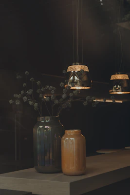 Two decorative vases, one dark green and the other orange, sit on a wooden table. Above them, three pendant lights with intricate designs cast a warm glow, creating a cozy and inviting atmosphere. Delicate branches with small, round seeds extend from the larger vase.