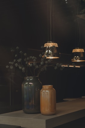 Two decorative vases, one dark green and the other orange, sit on a wooden table. Above them, three pendant lights with intricate designs cast a warm glow, creating a cozy and inviting atmosphere. Delicate branches with small, round seeds extend from the larger vase.