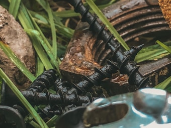 Close-up of several black screws lying on green grass and next to a rusted metal washer. Some stones are visible amidst the greenery, adding a natural texture to the scene.