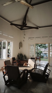 A cozy indoor space with wooden furniture arranged around a central table. Large windows allow natural light to fill the room, highlighting the plants and providing a view of a lush garden outside. The high ceiling is adorned with an industrial-style ceiling fan, and the room's aesthetic emphasizes natural materials and a connection to the outdoors.