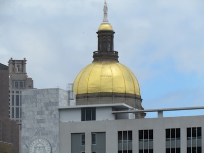 A prominent golden dome atop a government building is visible. The surrounding structures include modern and classical architecture, with a cloudy sky in the background.