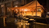 A cozy street scene at night featuring a coffee truck illuminated by warm, glowing lights. Two people sit at a small table under a canopy, enjoying drinks, while a barista works inside the van. The setup includes foldable chairs and tables, creating a casual, welcoming ambiance on a damp street with city buildings in the background.