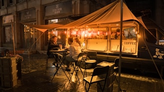 A cozy street scene at night featuring a coffee truck illuminated by warm, glowing lights. Two people sit at a small table under a canopy, enjoying drinks, while a barista works inside the van. The setup includes foldable chairs and tables, creating a casual, welcoming ambiance on a damp street with city buildings in the background.