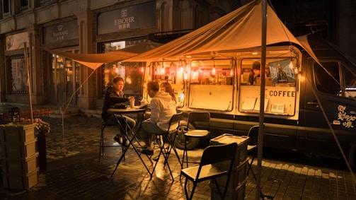 A cozy street scene at night featuring a coffee truck illuminated by warm, glowing lights. Two people sit at a small table under a canopy, enjoying drinks, while a barista works inside the van. The setup includes foldable chairs and tables, creating a casual, welcoming ambiance on a damp street with city buildings in the background.