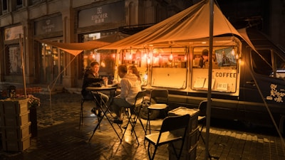 A cozy street scene at night featuring a coffee truck illuminated by warm, glowing lights. Two people sit at a small table under a canopy, enjoying drinks, while a barista works inside the van. The setup includes foldable chairs and tables, creating a casual, welcoming ambiance on a damp street with city buildings in the background.