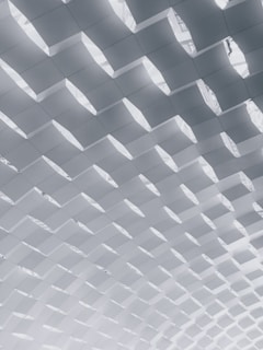 A geometric, repetitive pattern of angled, white panels creating a textured ceiling effect, with light filtering through the gaps.