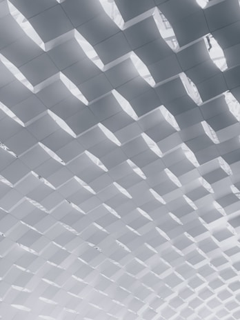 A geometric, repetitive pattern of angled, white panels creating a textured ceiling effect, with light filtering through the gaps.