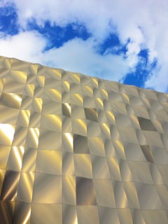 A modern building facade covered in reflective metallic panels arranged in a wavy pattern under a partly cloudy blue sky.