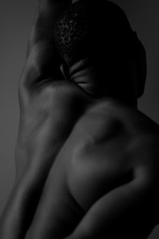 A black and white photograph captures the muscular back of a person with their arm raised, showcasing the contours and textures of the skin. The image focuses on the shadows and highlights, creating a dramatic effect.
