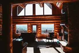 Warm sunlight streams through large windows in a rustic log cabin interior, revealing wooden beams and walls, and a cozy living space with a fireplace, armchair, and a couch. The view outside shows a sprawling landscape with forested hills.