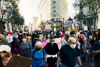 A large group of people is gathered in a city street, holding various protest signs advocating for women's rights and reproductive freedom. Many participants are wearing face masks, and the scene is set against a backdrop of tall buildings and trees.