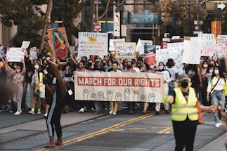A large group of people is participating in a protest march, holding various signs and banners. One prominent banner reads 'March for Our Rights' and features illustrations of raised fists. Many participants are wearing face masks, and some are wearing safety vests. The scene takes place on a city street, with buildings and trees visible in the background.