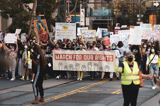 A large group of people is participating in a protest march, holding various signs and banners. One prominent banner reads 'March for Our Rights' and features illustrations of raised fists. Many participants are wearing face masks, and some are wearing safety vests. The scene takes place on a city street, with buildings and trees visible in the background.