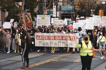 A large group of people is participating in a protest march, holding various signs and banners. One prominent banner reads 'March for Our Rights' and features illustrations of raised fists. Many participants are wearing face masks, and some are wearing safety vests. The scene takes place on a city street, with buildings and trees visible in the background.