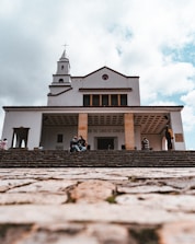 A large church building with a cross on top is viewed from the bottom of a stone staircase. The sky is partially covered with clouds. There are a few people sitting on the steps, some talking and others appearing contemplative.