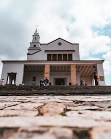 A large church building with a cross on top is viewed from the bottom of a stone staircase. The sky is partially covered with clouds. There are a few people sitting on the steps, some talking and others appearing contemplative.