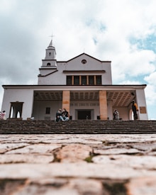 A large church building with a cross on top is viewed from the bottom of a stone staircase. The sky is partially covered with clouds. There are a few people sitting on the steps, some talking and others appearing contemplative.