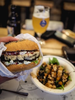 A close-up of a hand holding a gourmet burger wrapped in paper, featuring a variety of toppings such as crumbled cheese and vegetables. Beside it, a salad with greens, nuts, and croutons is served in a white bowl. A blurred background shows a glass of beer and a bottle on a wooden table.