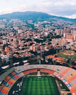 Aerial view of a large city with a prominent sports stadium in the foreground. The stadium features a soccer field and colorful seating sections. In the background, numerous tall buildings and residential areas spread across a hilly terrain, with lush greenery interspersed among the urban landscape.