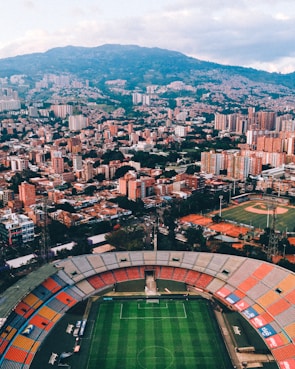 Aerial view of a large city with a prominent sports stadium in the foreground. The stadium features a soccer field and colorful seating sections. In the background, numerous tall buildings and residential areas spread across a hilly terrain, with lush greenery interspersed among the urban landscape.