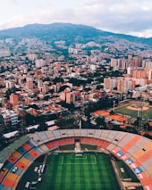 Aerial view of a large city with a prominent sports stadium in the foreground. The stadium features a soccer field and colorful seating sections. In the background, numerous tall buildings and residential areas spread across a hilly terrain, with lush greenery interspersed among the urban landscape.