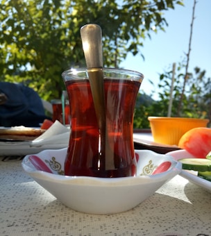 A glass of red tea in a traditional curved Turkish tea glass with a spoon inside, placed on a saucer with a decorative design. The setting is outdoors with sunlight filtering through green leaves in the background. There are additional food items nearby including slices of cucumber and tomato, along with an orange bowl.