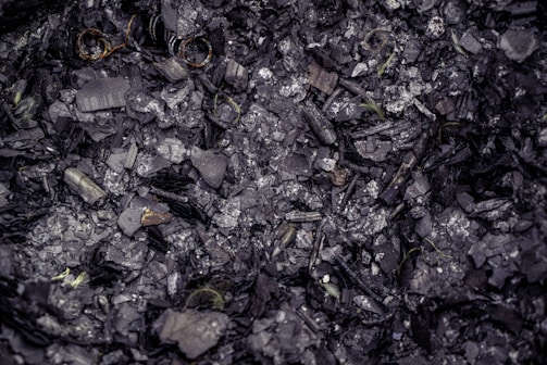 A mixture of charred debris and ash with small pieces of partially burned materials. The texture is rough and fragmented, and some areas show a metallic sheen.