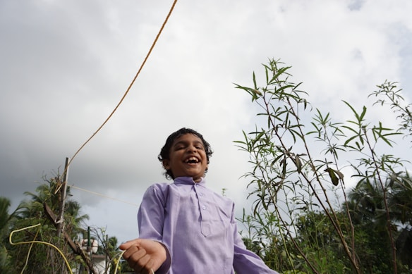 A joyful child is laughing outdoors, surrounded by lush green plants under a cloudy sky. The child is wearing a light purple shirt and appears to be in a garden or natural setting.