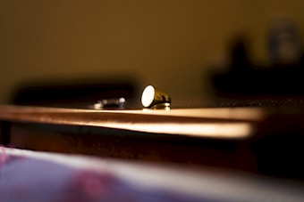 A close-up shot of a single brass-colored bullet casing lying on a wooden surface. The background is slightly out of focus, giving the image a dramatic and intense feel. Light gently illuminates the surface and the casing, creating a warm, moody atmosphere.
