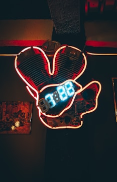 A neon-lit sign featuring a stylized engine with glowing blue digital numbers at its center. The backdrop is dark, making the red and blue neon tones of the engine stand out prominently. The setting appears to be indoors, with a framed picture visible on the left side.
