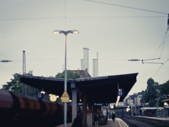 A train station platform with overhead lights and a view of industrial buildings with tall chimneys in the background. The platform is labeled with 'B' signs, and a train is stationed on the left side. Overhead wires stretch across the sky, and a few people are waiting on the platform, with the station extending into the distance.
