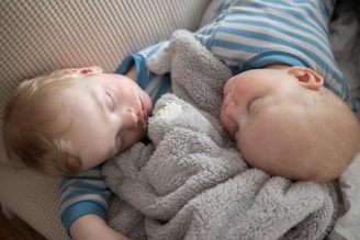 Two young children are peacefully sleeping side by side, lying on a comfortable-looking surface with soft textures. They are both wearing blue and white striped pajamas. A plush, gray blanket is nestled between them, adding to the cozy atmosphere.