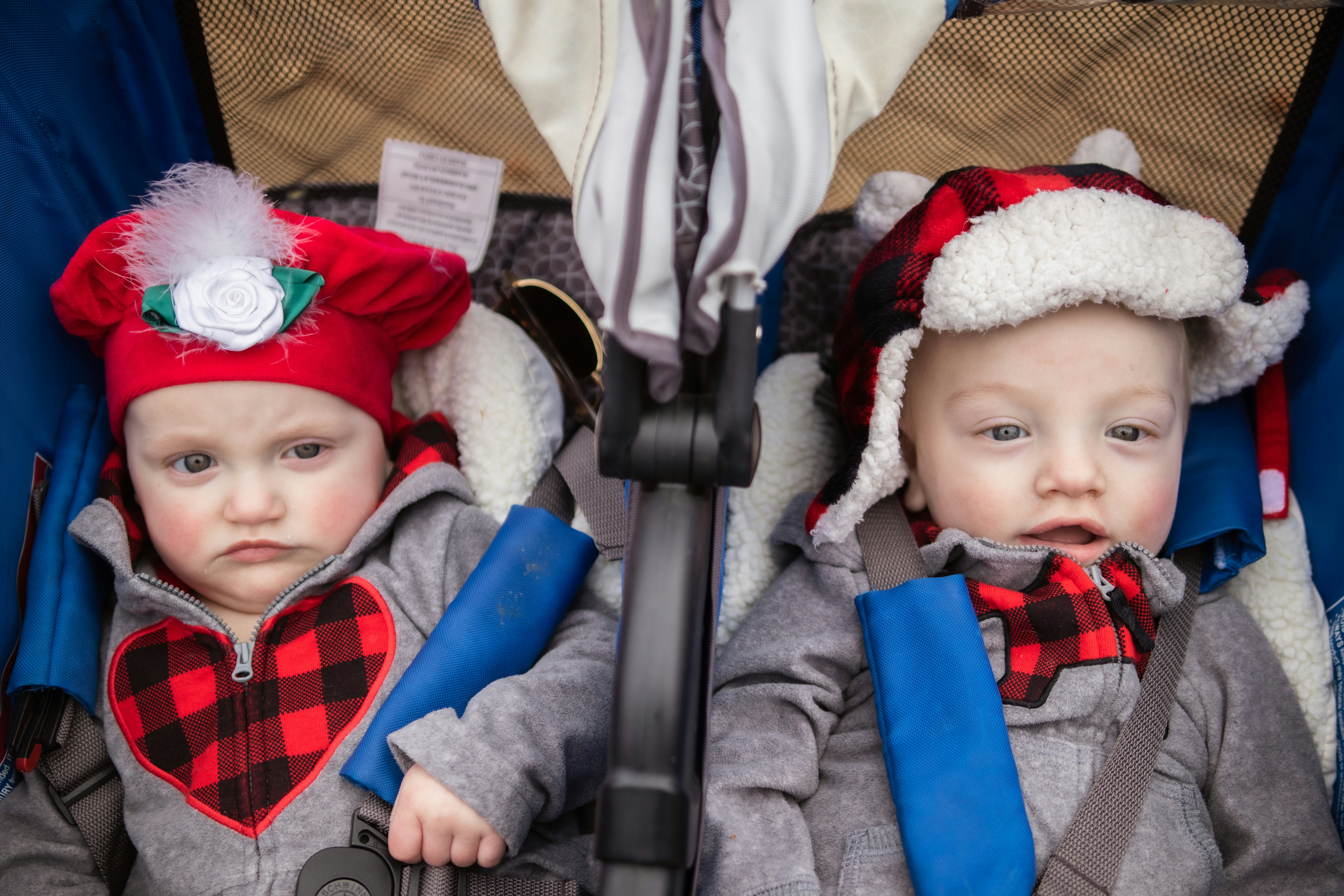 Two babies side by side in a double stroller, each buckled into a harness with padded shoulder straps