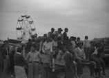 A group of people, including men, women, and children, are gathered around and sitting on a wooden cart or wagon, possibly during a community event or fair. In the background, a Ferris wheel is partly visible, indicating a fair or celebration. Most people appear to be dressed in casual clothes, suitable for an outdoor event. The atmosphere seems lively and social.