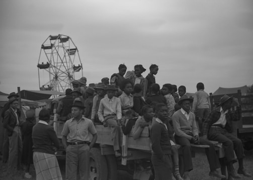 A group of people, including men, women, and children, are gathered around and sitting on a wooden cart or wagon, possibly during a community event or fair. In the background, a Ferris wheel is partly visible, indicating a fair or celebration. Most people appear to be dressed in casual clothes, suitable for an outdoor event. The atmosphere seems lively and social.