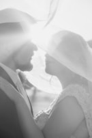 A black and white image captures a couple embracing under a sheer veil, with light streaming in from the background. The scene conveys a sense of intimacy and tenderness, enhanced by the soft focus and gentle textures of their attire.