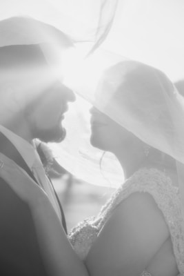 A black and white image captures a couple embracing under a sheer veil, with light streaming in from the background. The scene conveys a sense of intimacy and tenderness, enhanced by the soft focus and gentle textures of their attire.