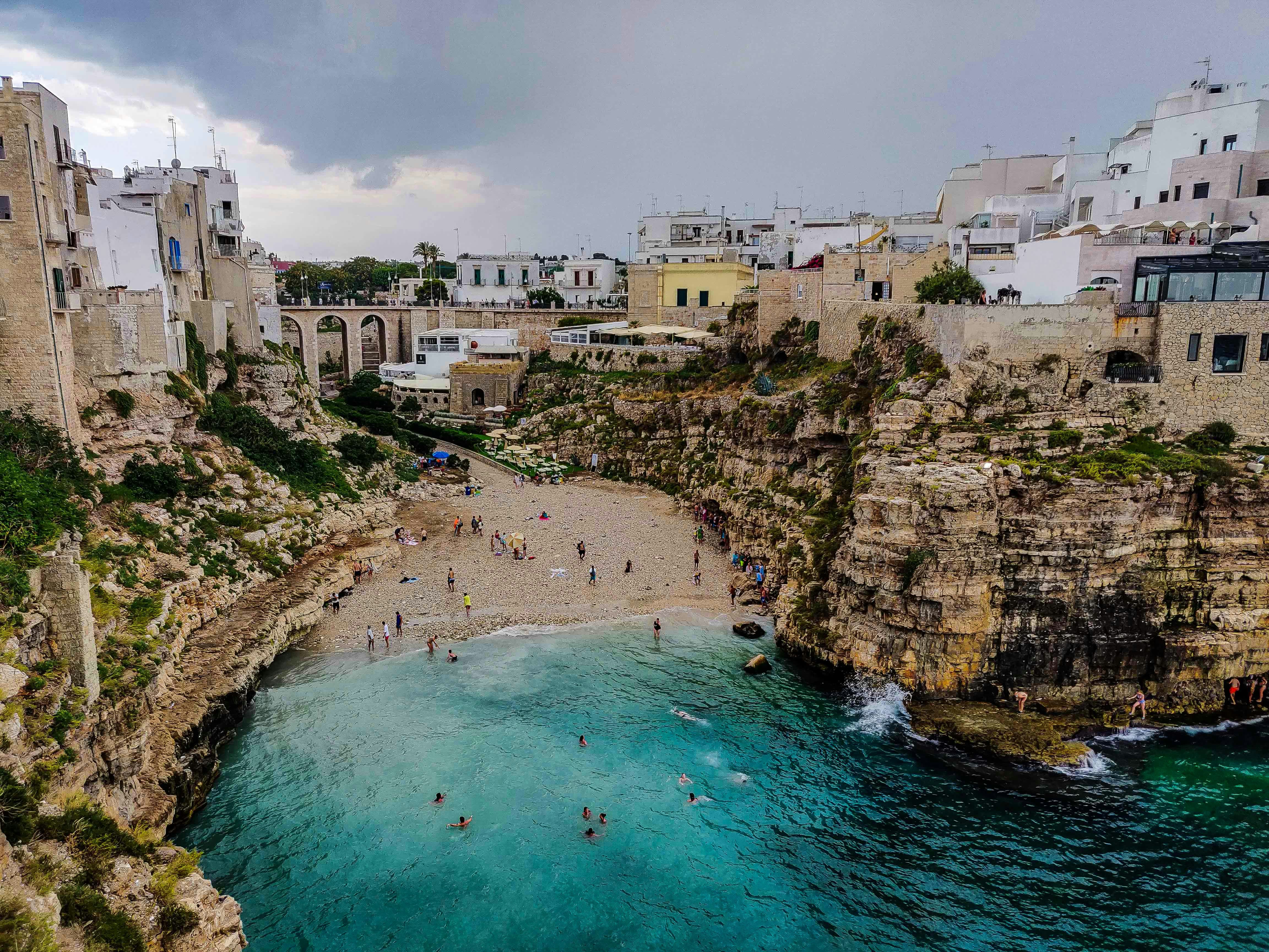 a group of people standing on top of a cliff next to a body of water, A cloudy day on an Italian beach