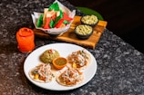 A delicious spread of Mexican cuisine featuring three tacos topped with shredded vegetables and sauce, a serving of green rice with a tomato garnish, a bowl of green salsa, and colorful red, green, and beige tortilla chips. Two bowls of guacamole are placed on a wooden tray, and a vibrant red-orange drink sits next to the plate.