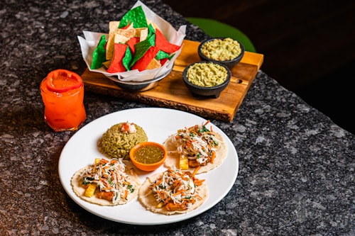 A delicious spread of Mexican cuisine featuring three tacos topped with shredded vegetables and sauce, a serving of green rice with a tomato garnish, a bowl of green salsa, and colorful red, green, and beige tortilla chips. Two bowls of guacamole are placed on a wooden tray, and a vibrant red-orange drink sits next to the plate.