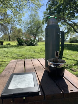A tranquil outdoor setting features a picnic table with a digital e-reader displaying text, a large metallic thermos, and a traditional mate cup. Sunlight filters through the surrounding trees, casting shadows on the table. The background consists of lush green grass and a variety of trees, suggesting a park or garden environment.