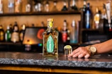 A bottle of Hornitos tequila is placed on a bar counter alongside a shot glass filled with tequila and a slice of lime on top. A hand wearing a gold watch rests on the counter near the shot glass. The background consists of a variety of blurred bottles and bar shelves.