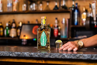 A bottle of Hornitos tequila is placed on a bar counter alongside a shot glass filled with tequila and a slice of lime on top. A hand wearing a gold watch rests on the counter near the shot glass. The background consists of a variety of blurred bottles and bar shelves.
