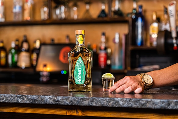 A bottle of Hornitos tequila is placed on a bar counter alongside a shot glass filled with tequila and a slice of lime on top. A hand wearing a gold watch rests on the counter near the shot glass. The background consists of a variety of blurred bottles and bar shelves.